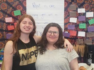 Two white women stand in front of a colorful tapestry