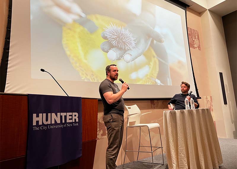 Caption: Andrew Robinson (left) and his partner Dominic Smith (right) shared their experience of documenting carnivorous purple sea urchins (shown on screen) by diving deep into waters of Monterey Bay. According to their story, rising population of sea urchins has significantly diminished kelp forests. [Photo by Professor Sissel McCarthy]